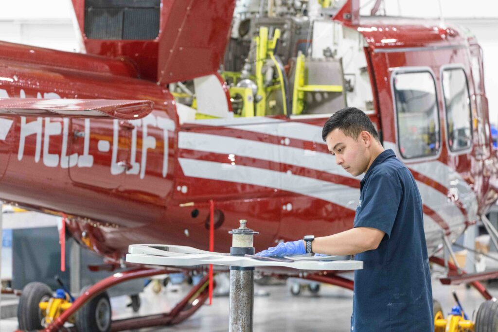 Technician working on a rotor blade beside a red Heli-Lift helicopter in a hangar at Villeneuve Airport